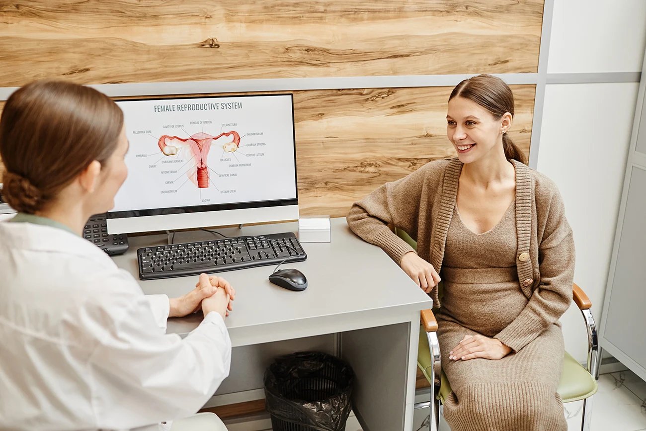 Pregnant woman smiling during a consultation with a gynecologist showing a female reproductive system diagram.